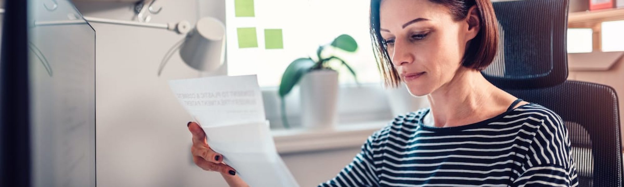 Woman sitting by the window and reading mail at the office Woman wearing striped shirt sitting by the window at the table and reading mail in the office